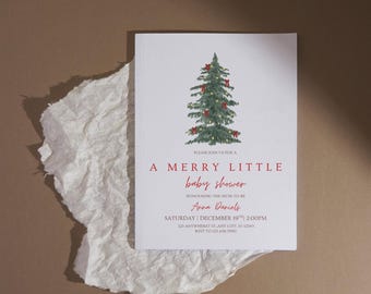 May include: A white invitation card with a Christmas tree illustration and red text. The text reads "A Merry Little Baby Shower" with event details. The card is on a brown surface with a piece of torn white paper.