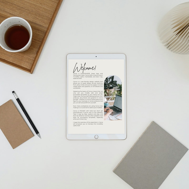 May include: A flat lay featuring a tablet displaying the word "Welcome" and text about Canva. A cup of tea, a notebook, a pencil, and a decorative fan are also present. The scene is set on a white surface, creating a clean and organized workspace.