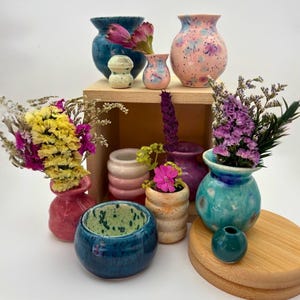May include: A selection of vibrant ceramic vases in different sizes and shapes, some containing dried flowers. The vases are in shades of blue, pink, purple, and green, with speckled and glazed finishes. Some are cylindrical, others rounded.