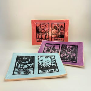 May include: Three rectangular ceramic trays in coral, lavender, and light blue. Each tray features black illustrations of tarot cards, including "The Moon" and "The Sun." The trays have a light tan rim.