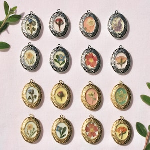May include: An assortment of oval-shaped lockets with pressed flowers inside. The lockets have either silver or gold-colored frames. Green leafy sprigs are placed on the top and bottom of the image, with a light pink background.