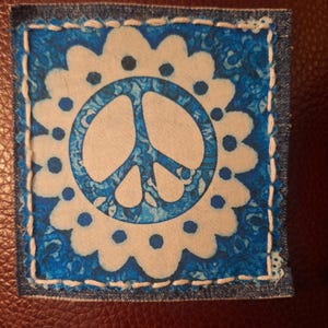 May include: Square patch with a peace sign design. The patch features a white peace symbol in a blue circle, surrounded by a white floral design on a blue patterned background. The patch is bordered by denim and white stitching.