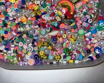 May include: A clear plastic container filled with colorful plastic beads. The beads are in various shapes, including circles, squares, and stars. There are also some beads with letters and smiley faces on them.