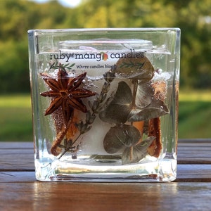 May include: A square glass jar candle with a white candle inside. The jar contains dried botanicals, including star anise, eucalyptus leaves, and cinnamon sticks. The jar has a label that reads "ruby mango candles" and "where candles bloom".