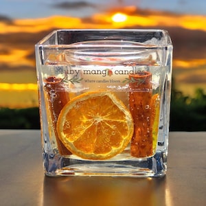 Op de afbeelding: Een heldere glazen pot met een kaars, een gedroogde sinaasappelschijf en kaneelstokjes. De kaars is gelabeld met "ruby mango candles" en "Where candles bloom".