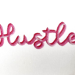 May include: Pink embroidered word "Hustle" on a white fabric background.