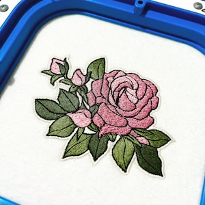 Può includere: Un ricamo che raffigura una grande rosa rosa in piena fioritura, accompagnata da diversi boccioli di rosa più piccoli e foglie verde scuro. Il disegno è cucito su una base di tessuto bianco fissata all'interno di un telaio da ricamo blu.