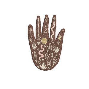 Pode incluir: Uma mão marrom com detalhes bordados, incluindo uma lua crescente, estrelas, um cacto, uma cobra e a palavra "paz".