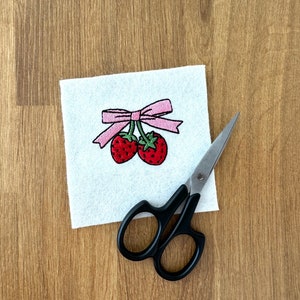 May include: A square piece of white felt featuring an embroidered design of two red strawberries with green stems tied together by a light pink ribbon bow, positioned next to a pair of black handled scissors with silver metal blades.