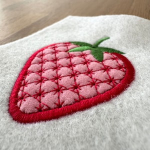 May include: An embroidered strawberry patch on a white felt background. The strawberry features a light pink quilted center with dark red cross-stitch detailing, a solid red embroidered border, and a green embroidered stem at the top.