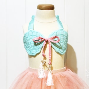 Könnte beinhalten: Ein hellblaues Stoff-Muschel-Oberteil mit einem rosa gepunkteten Band, das zu einer Schleife gebunden ist. Das Oberteil befindet sich auf einem weißen Mannequin und hat weiße Quasten, die daran hängen.