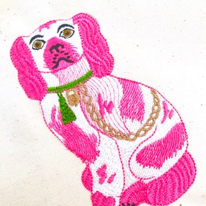May include: Embroidered dog design in pink and white, featuring a green collar and gold chain. The dog has brown eyes and a black nose. The embroidery is on a cream-colored fabric.