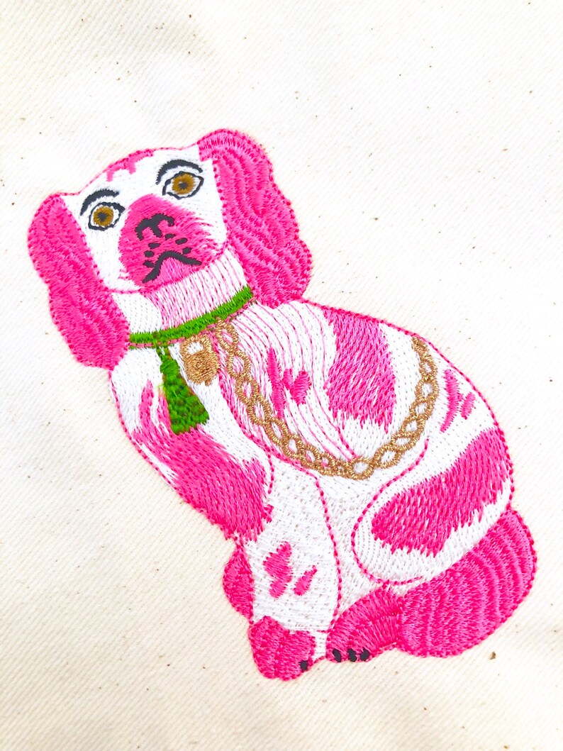 May include: Embroidered dog design in pink and white, featuring a green collar and gold chain. The dog has brown eyes and a black nose. The embroidery is on a cream-colored fabric.