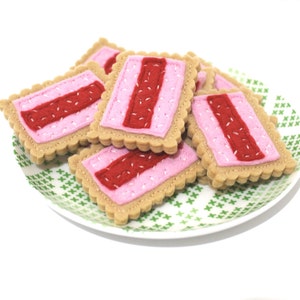 May include: A plate of felt cookies with pink and red frosting. The cookies are shaped like rectangles and have a scalloped edge.