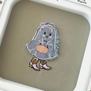 Può includere: Un fantasma di denim blu con un fiocco rosa che tiene una piccola zucca arancione. Il fantasma ha un viso amichevole con occhi neri e un blush rosa. Il fantasma sta in piedi su gambe marroni con scarpe rosa.
