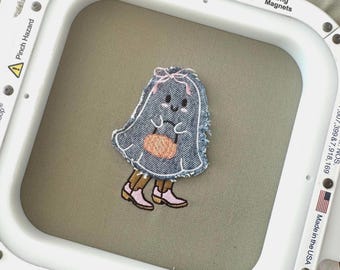 Puede incluir: Un fantasma de mezclilla azul con un lazo rosa sosteniendo una pequeña calabaza naranja. El fantasma tiene una cara amigable con ojos negros y un rubor rosado. El fantasma está de pie sobre piernas marrones con zapatos rosas.