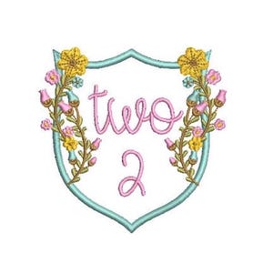 May include: An embroidery design featuring a light blue shield outline containing the pink text two and 2. The shield is framed by floral vines with yellow flowers, pink buds, and green stems, creating a symmetrical decorative emblem.