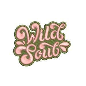 Op de afbeelding: Een geborduurde patch met de tekst Wild Soul in een cursief, lichtroze lettertype met een dikke, donker olijfgroene rand. Het borduurwerk heeft een zichtbare stikseltextuur door het hele ontwerp, wat een verhoogd uiterlijk geeft.