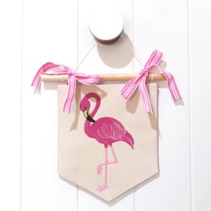 May include: A pink flamingo embroidered on a beige fabric banner with pink and white striped ribbon hanging from a wooden dowel.