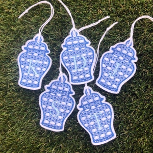 May include: Five blue and white porcelain ginger jar ornaments with a light blue monogram on each. The ornaments are hanging from white string.