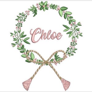 May include: An embroidery design featuring a circular wreath of green leaves and small pink flowers. The name Chloe is stitched in pink script in the center. At the bottom, a tan rope-like bow with two dangling pink tassels completes the design.