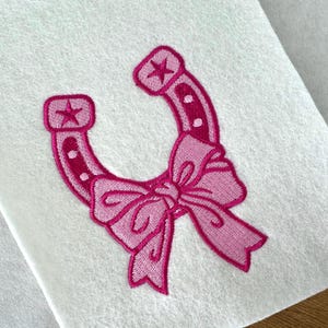 May include: An embroidered patch on white fabric featuring a bright pink horseshoe with a dark pink outline. The horseshoe has a star at each end and is decorated with a large, light pink bow tied at the bottom center with dark pink stitching.