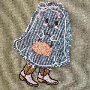 Può includere: Un fantasma di jeans blu con un fiocco rosa e una zucca rosa in mano. Il fantasma indossa stivali marroni con accenti rosa.