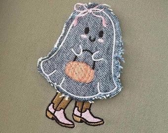 Puede incluir: Un fantasma de mezclilla azul con un lazo rosa y una calabaza rosa en la mano. El fantasma lleva botas marrones con detalles rosas.