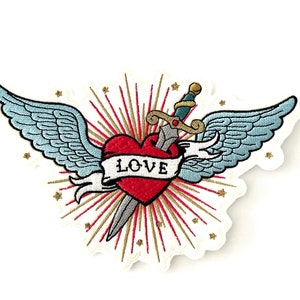 May include: A red heart with wings and a dagger through it, surrounded by a gold and red starburst. The heart has the word "LOVE" written on it.