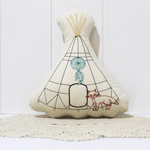 May include: A cream-colored teepee-shaped pillow with embroidered details. The design includes a dreamcatcher, geometric lines, and a fox. The pillow is placed on a lace doily, creating a rustic, bohemian aesthetic.