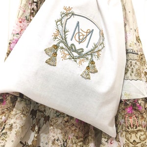 May include: A white fabric bag with a monogram embroidered on it. The monogram is a stylized "M" and "M" with a floral design around it. The bag is lying on a floral fabric.