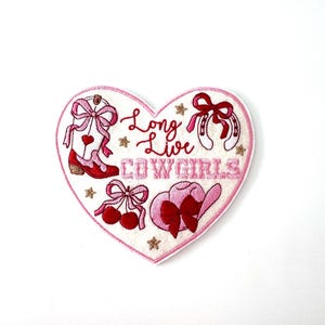 May include: A heart-shaped embroidered patch featuring a white background with red and pink designs including a cowboy boot, a horseshoe, cherries, and a hat. The text reads Long Live COWGIRLS in red and pink, accented by small gold stars.