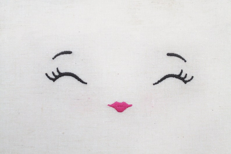 May include: Embroidered white fabric with a simple face design. The face has two black eyelashes and a pink smiling mouth.