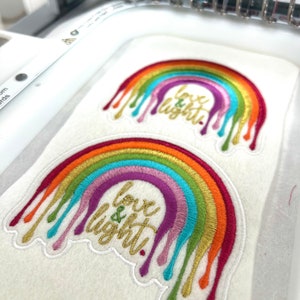 Peut inclure: Deux écussons brodés sur tissu blanc présentant un motif arc-en-ciel avec des effets de peinture dégoulinante en rouge, orange, jaune, vert, bleu et violet. Le texte "love & light" est brodé en fil doré au centre de chaque arc-en-ciel.