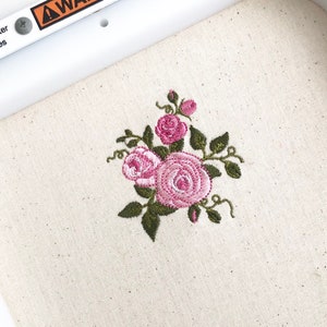 May include: An embroidery design featuring a cluster of three pink roses with dark green leaves and stems stitched onto a light beige fabric. A white plastic frame edge with a partial orange warning label is visible at the top of the image.