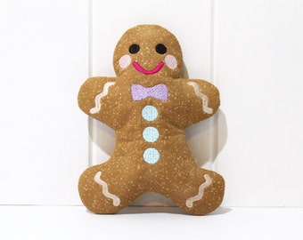 May include: A gingerbread man plush toy with a brown body, white icing details, and a purple bow tie. The toy has a smiling face with black eyes, pink cheeks, and a pink mouth.