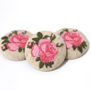 Puede incluir: Tres botones redondos de tela beige con rosas bordadas de color rosa y hojas verdes.