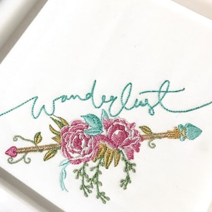 Puede incluir: Tela blanca con un diseño bordado con la palabra "Wanderlust" en escritura turquesa sobre una flecha floral. La flecha tiene flores rosas y amarillas, hojas turquesas y un asta dorada.