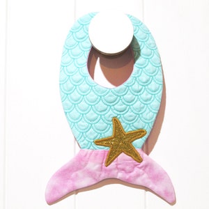 May include: A turquoise and pink mermaid-shaped baby bib with a gold glitter starfish.