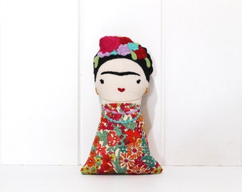 May include: A handmade doll resembling Frida Kahlo, featuring a cream-colored face with black eyebrows, a red mouth, and a floral headdress. The doll wears a colorful floral dress with a turquoise necklace.