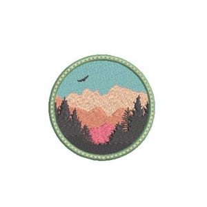 May include: A circular embroidered patch featuring a landscape scene with light blue sky, a small black bird in flight, tan and pink mountain peaks, and dark grey evergreen trees in the foreground, all enclosed within a light green stitched border.