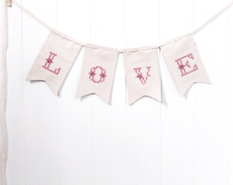 Puede incluir: Una pancarta de tela blanca con la palabra "LOVE" escrita en letras bordadas de color rosa. Cada letra está en una bandera separada con la parte inferior puntiaguda.