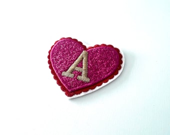 Può includere: Un cuore di feltro rosa con un bordo a festoni e una lettera A ricamata in oro al centro.