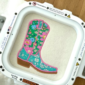 May include: A white embroidery hoop with a floral patterned boot design. The boot is pink, green, and blue with a brown sole. The hoop is labelled "6.5" Mighty Hoop" and has a warning label that says "Pinch Hazard".