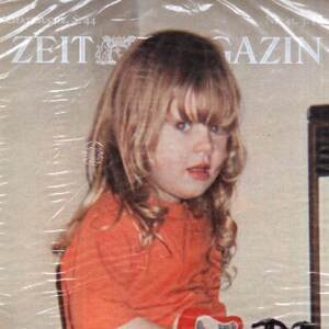 May include: A magazine cover featuring a young child in an orange shirt and blue jeans. The child is holding a red toy guitar. The magazine title is "ZEIT MAGAZIN". The cover is wrapped in plastic.
