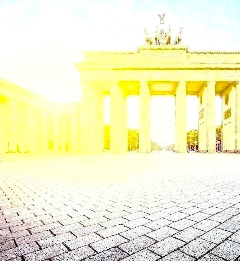 May include: A sunlit view of the Brandenburg Gate in Berlin, Germany. The neoclassical structure has tall columns and a quadriga sculpture. The foreground shows a grey brick pathway.