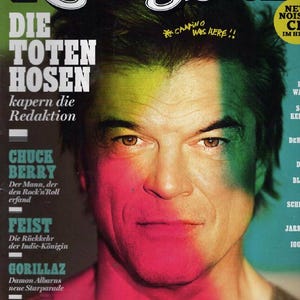 Könnte beinhalten: Ein Rolling Stone Magazin-Cover mit dem Porträt einer Person mit dunklem Haar, beleuchtet durch grünes und pinkes Licht. Der Text umfasst 'Rolling Stone', 'DIE TOTEN HOSEN', 'CHUCK BERRY', 'FEIST', 'GORILLAZ', 'ANARCHOS IM KRIEG' und 'CAMINO WAS HERE!!'.