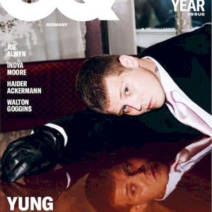 Könnte beinhalten: GQ Deutschland Magazin-Cover mit Yung Lean. Das Cover enthält den Text "The 2025 Men of the Year Issue". Das Bild zeigt einen Mann in Anzug und Krawatte mit schwarzen Handschuhen, der sich auf eine reflektierende Oberfläche stützt.