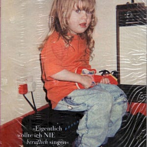 Könnte beinhalten: Ein Magazin-Cover mit dem Titel ZEITmagazin, das ein Kind mit langem blonden Haar in einem orangefarbenen T-Shirt und einer hellblauen Acid-Wash-Jeans zeigt. Das Kind hält eine kleine rote Spielzeuggitarre. Der Text lautet: Eigentlich wollte ich NIE beruflich singen.