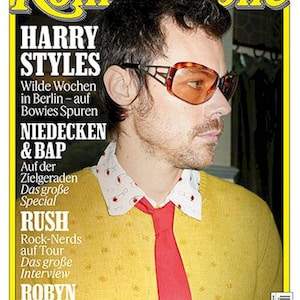 May include: Rolling Stone magazine cover with Harry Styles. He is wearing orange sunglasses, a yellow sweater, a red tie, and a patterned collared shirt. The magazine's masthead and cover lines are visible, including "April 2026" and "Germany."